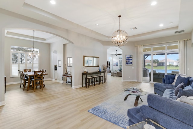 living room with a healthy amount of sunlight, ceiling fan with notable chandelier, a tray ceiling, and light hardwood / wood-style floors