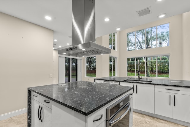 kitchen featuring island range hood, white cabinetry, sink, dark stone counters, and a center island