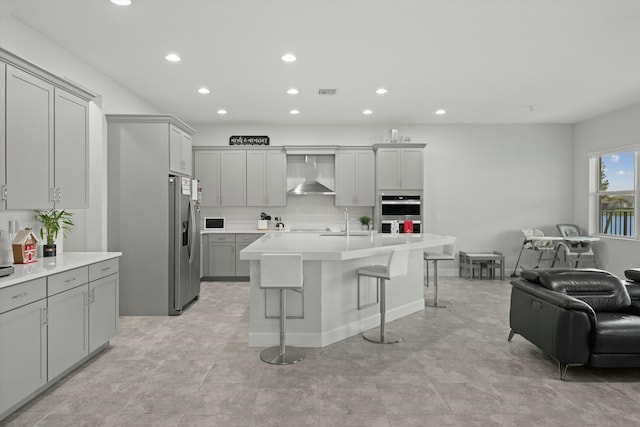 kitchen with gray cabinetry, light countertops, wall chimney range hood, an island with sink, and a kitchen bar