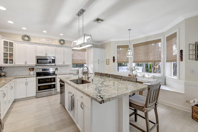 kitchen featuring a kitchen island with sink, sink, appliances with stainless steel finishes, decorative light fixtures, and white cabinetry