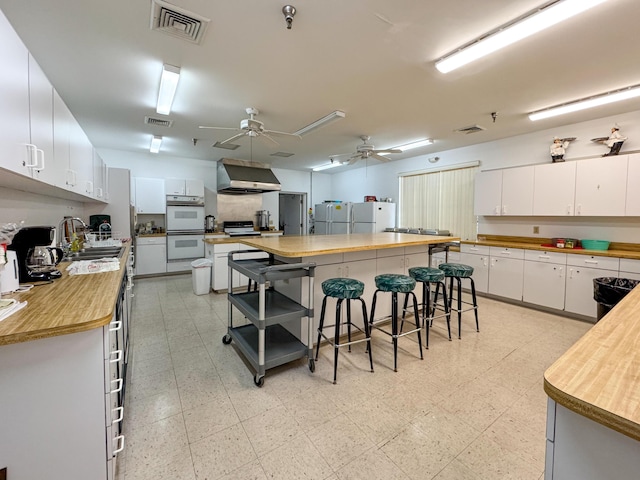 kitchen featuring wall chimney range hood, sink, white appliances, white cabinetry, and a breakfast bar area