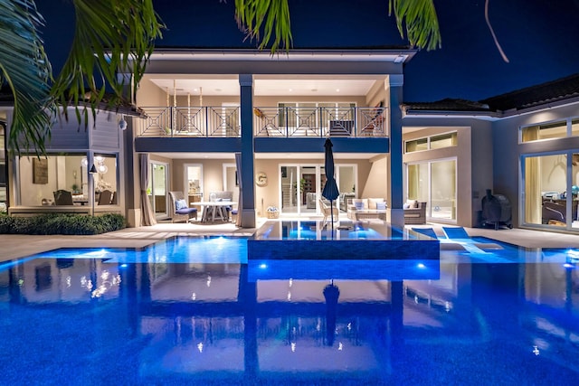 back house at night with a pool with hot tub, a patio area, a balcony, and french doors