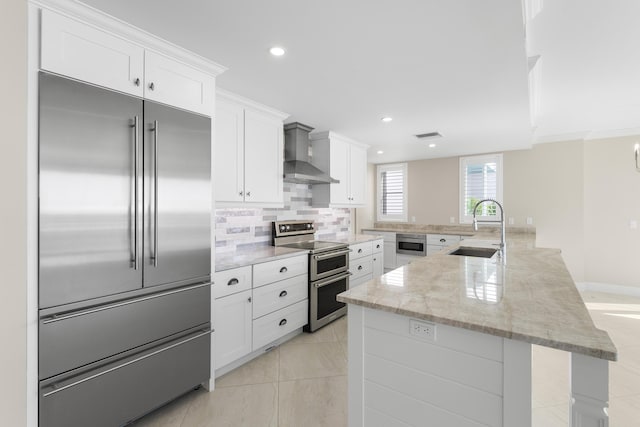 kitchen with white cabinets, sink, built in appliances, wall chimney exhaust hood, and light stone countertops