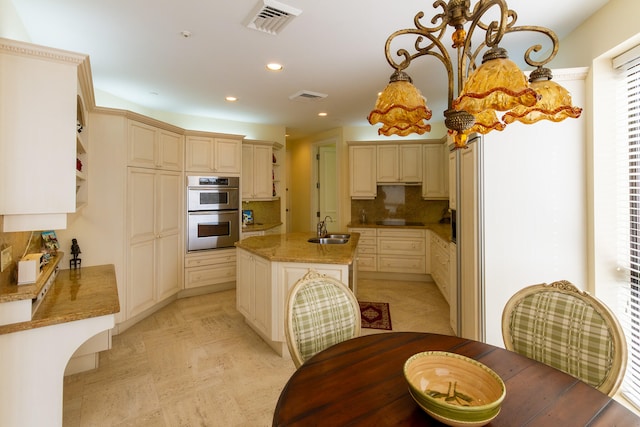 kitchen with sink, a kitchen island with sink, stainless steel double oven, cream cabinetry, and decorative backsplash