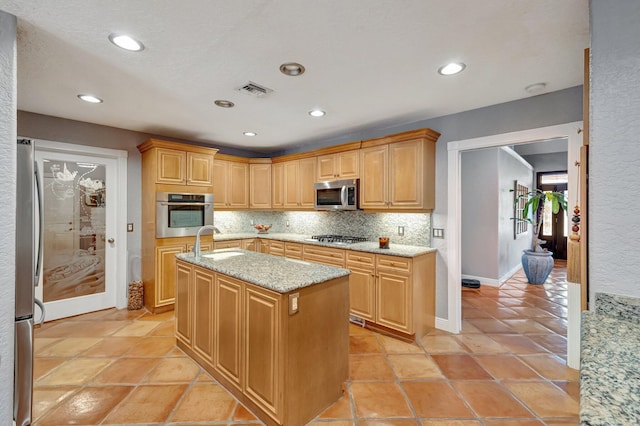 kitchen with light brown cabinets, light stone countertops, stainless steel appliances, and an island with sink