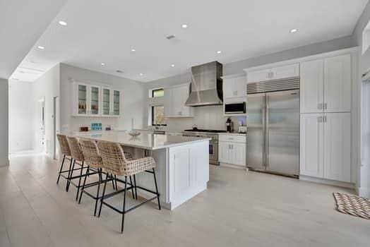 kitchen featuring a kitchen bar, a kitchen island with sink, stainless steel appliances, white cabinets, and wall chimney exhaust hood