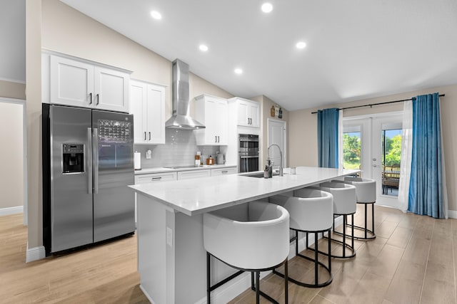 kitchen with appliances with stainless steel finishes, wall chimney exhaust hood, white cabinets, lofted ceiling, and an island with sink