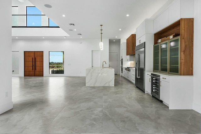 kitchen with white cabinets, built in refrigerator, wine cooler, hanging light fixtures, and an island with sink