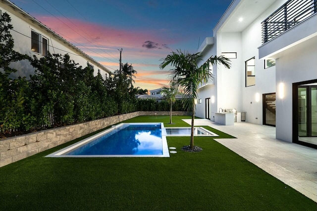 pool at dusk featuring a lawn, an outdoor kitchen, and a patio area