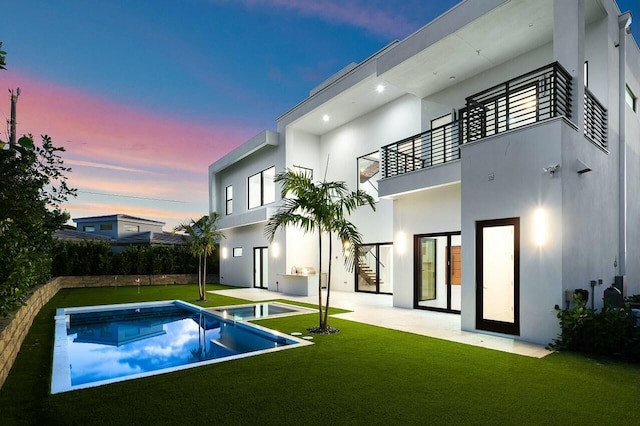 back house at dusk featuring a swimming pool with hot tub, a patio, a balcony, and a lawn