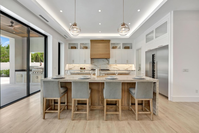 kitchen featuring stainless steel built in refrigerator, premium range hood, an island with sink, decorative light fixtures, and white cabinets