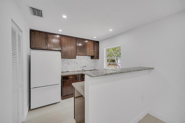 kitchen with white refrigerator, kitchen peninsula, decorative backsplash, light stone countertops, and dark brown cabinetry