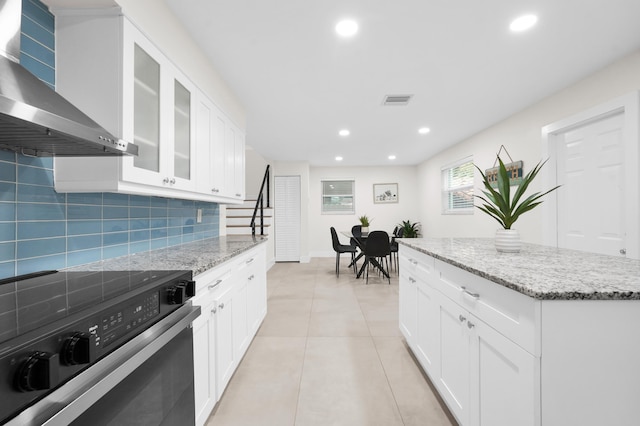 kitchen featuring decorative backsplash, light stone counters, wall chimney range hood, white cabinets, and stainless steel electric range