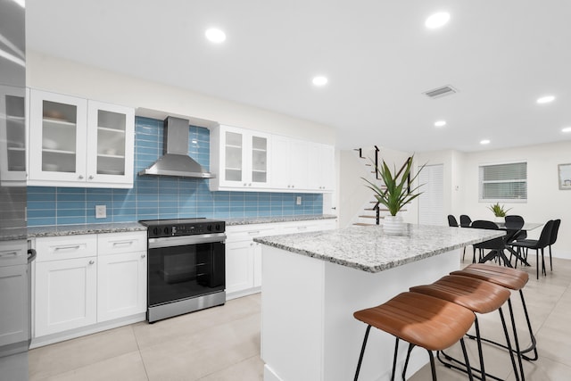 kitchen with electric stove, white cabinetry, a center island with sink, and wall chimney range hood