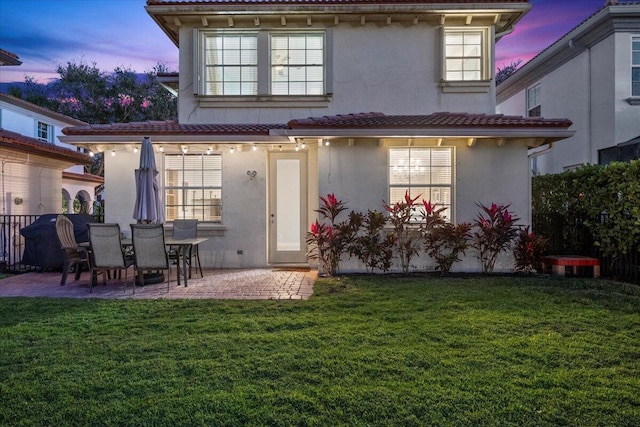 back house at dusk featuring a lawn and a patio area