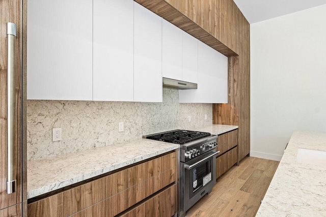 kitchen with under cabinet range hood, white cabinets, tasteful backsplash, high end range, and light wood finished floors