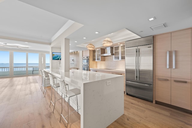 kitchen featuring a spacious island, a water view, wall chimney range hood, appliances with stainless steel finishes, and a kitchen bar