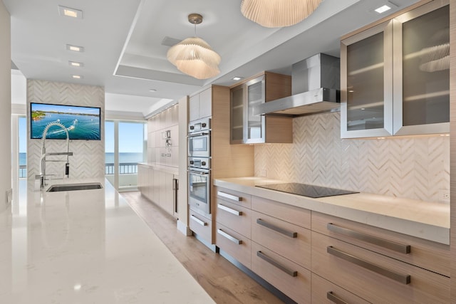 kitchen featuring light stone countertops, wall chimney exhaust hood, black electric cooktop, sink, and hanging light fixtures