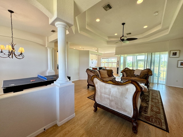 living room with ceiling fan with notable chandelier, a tray ceiling, ornate columns, and light hardwood / wood-style floors