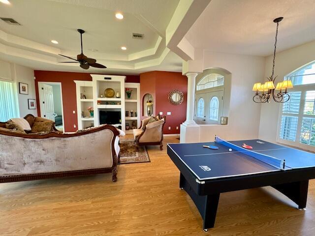 rec room with ceiling fan with notable chandelier, a raised ceiling, decorative columns, and light hardwood / wood-style flooring