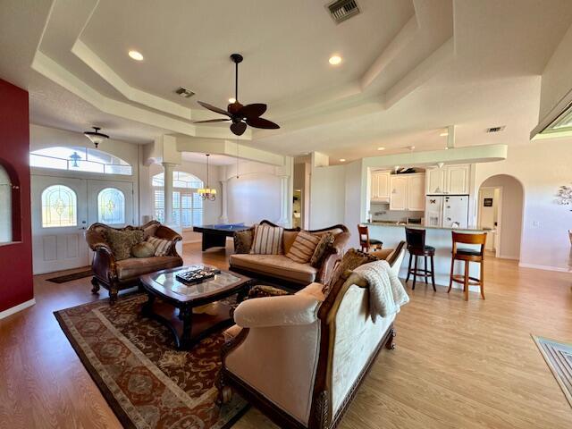 living room featuring ceiling fan with notable chandelier, a raised ceiling, french doors, and light hardwood / wood-style flooring