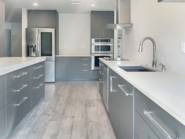 kitchen with gray cabinetry, wall chimney range hood, sink, appliances with stainless steel finishes, and light hardwood / wood-style floors