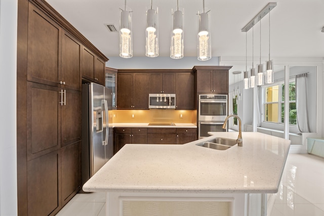 kitchen with a center island with sink, a sink, stainless steel appliances, dark brown cabinets, and hanging light fixtures