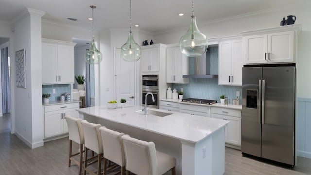 kitchen with light countertops, appliances with stainless steel finishes, a kitchen island with sink, a sink, and wall chimney exhaust hood