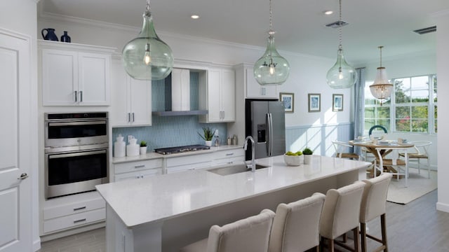 kitchen with sink, stainless steel appliances, wall chimney range hood, an island with sink, and white cabinets