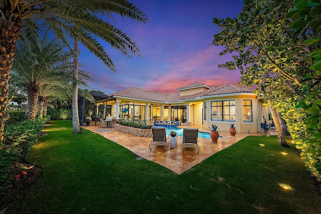 back house at dusk featuring a lawn and a patio area