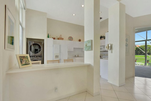 kitchen with kitchen peninsula, white appliances, white cabinets, stacked washer and dryer, and light tile patterned flooring