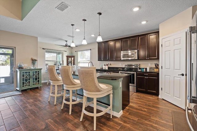 kitchen with ceiling fan, stainless steel appliances, an island with sink, decorative light fixtures, and a breakfast bar