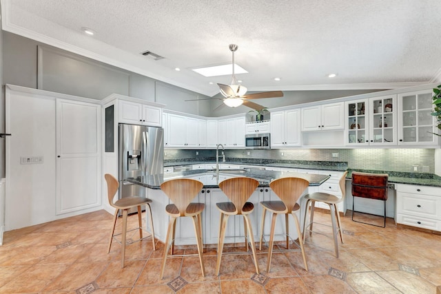 kitchen featuring appliances with stainless steel finishes, dark stone counters, vaulted ceiling with skylight, a kitchen island with sink, and white cabinetry