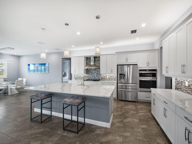 kitchen with appliances with stainless steel finishes, a kitchen island with sink, wall chimney range hood, pendant lighting, and white cabinetry