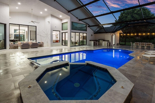 pool at dusk featuring glass enclosure, ceiling fan, an outdoor kitchen, an in ground hot tub, and a patio