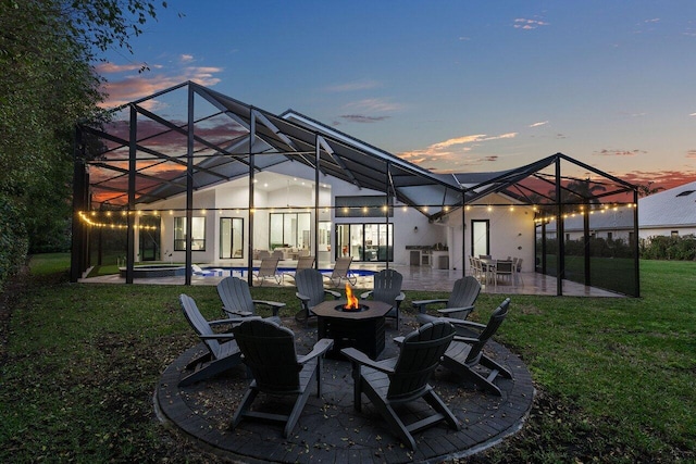 back house at dusk featuring a yard, a patio, glass enclosure, and an outdoor fire pit