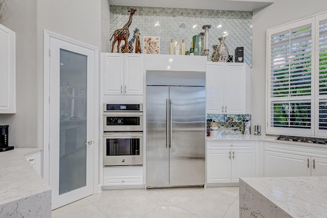 kitchen with appliances with stainless steel finishes, white cabinetry, decorative backsplash, light tile patterned flooring, and light stone counters