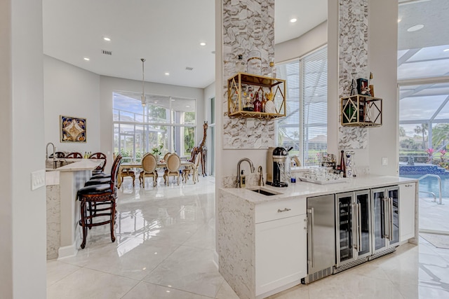 bar featuring beverage cooler, white cabinetry, sink, hanging light fixtures, and light stone counters