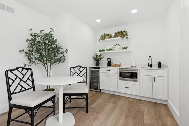 bar featuring stainless steel microwave, white cabinets, sink, wine cooler, and light hardwood / wood-style floors