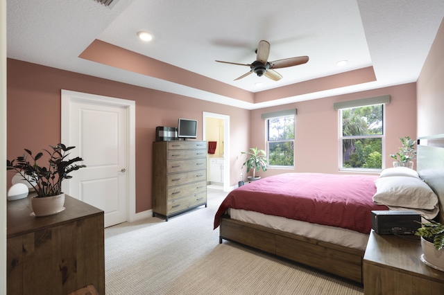 carpeted bedroom featuring a raised ceiling, ensuite bath, and ceiling fan