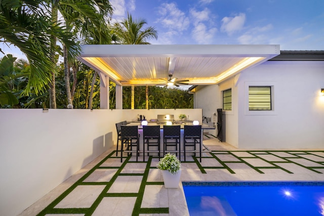 patio terrace at dusk with a fenced in pool, exterior bar, ceiling fan, a grill, and exterior kitchen