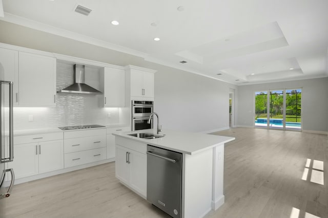 kitchen featuring stainless steel appliances, a raised ceiling, wall chimney range hood, white cabinets, and an island with sink