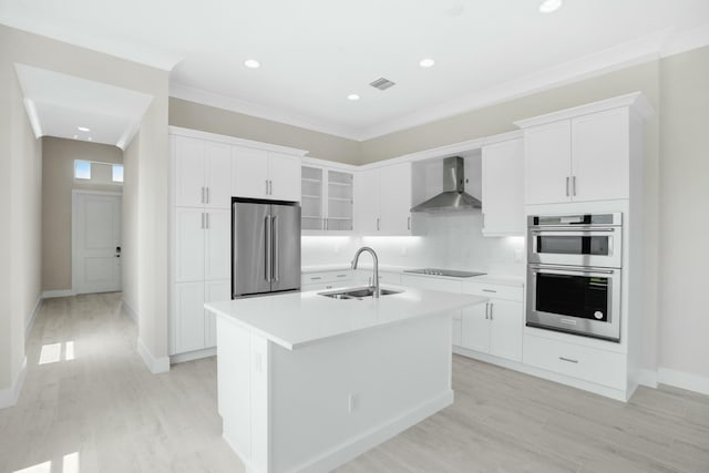 kitchen with wall chimney range hood, sink, an island with sink, appliances with stainless steel finishes, and white cabinetry