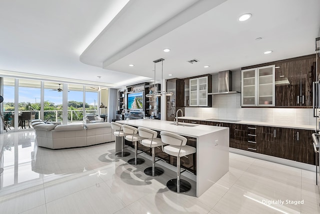 kitchen featuring backsplash, wall chimney exhaust hood, a kitchen island with sink, light tile patterned floors, and a breakfast bar area