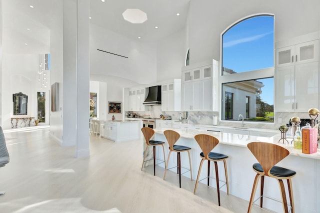 kitchen with wall chimney range hood, sink, a towering ceiling, tasteful backsplash, and white cabinetry