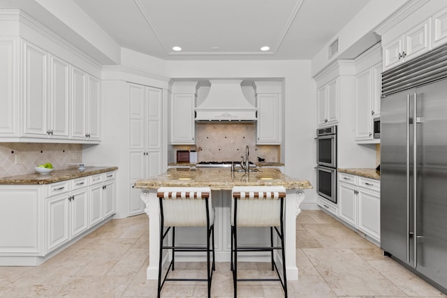 kitchen with custom range hood, stainless steel appliances, white cabinetry, and a center island with sink