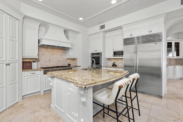 kitchen with built in appliances, decorative backsplash, a center island with sink, white cabinets, and custom exhaust hood