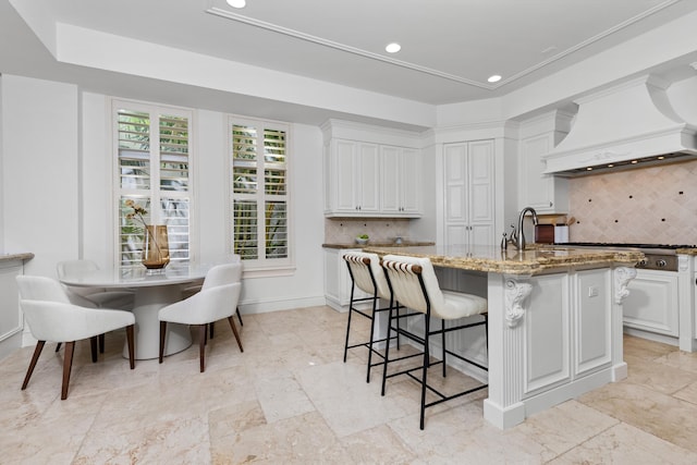 kitchen featuring a center island with sink, decorative backsplash, custom range hood, light stone counters, and white cabinetry