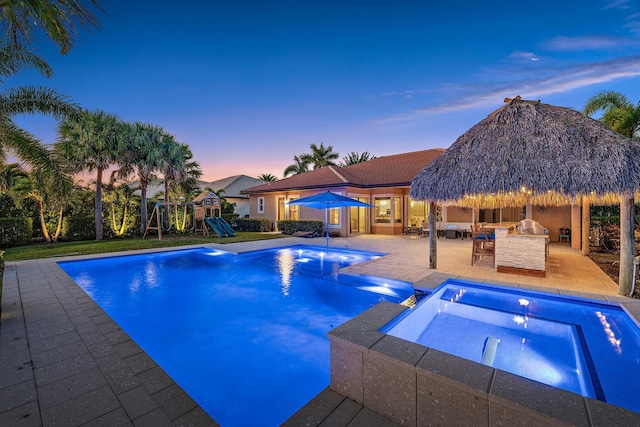 pool at dusk featuring an outdoor kitchen, a playground, an in ground hot tub, and a patio