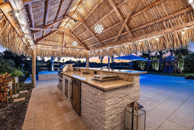 patio terrace at dusk featuring a gazebo, a playground, area for grilling, a pool, and exterior kitchen